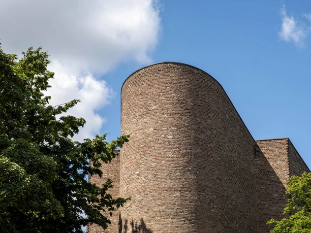 St. Alban Backsteinfassade einer modernen Kirche, teilweise von grünen Bäumen eingerahmt, unter blauem Himmel.