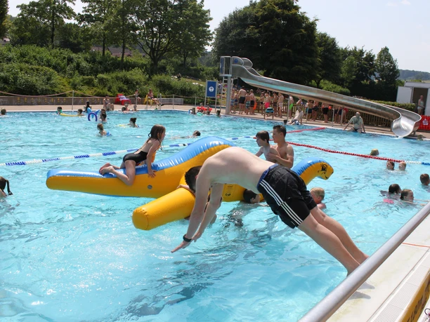 IMG_7701.JPG Ein belebtes Freibad im Sommer mit Badegästen auf einer aufblasbaren Wasserrutsche und Schwimmern.