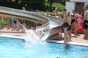 IMG_7748.JPG Eine Person rutscht mit Freude von einer Wasserrutsche in ein belebtes, sonniges Schwimmbad.