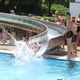 IMG_7748.JPG Eine Person rutscht mit Freude von einer Wasserrutsche in ein belebtes, sonniges Schwimmbad.