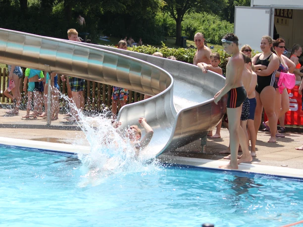 IMG_7748.JPG Eine Person rutscht mit Freude von einer Wasserrutsche in ein belebtes, sonniges Schwimmbad.