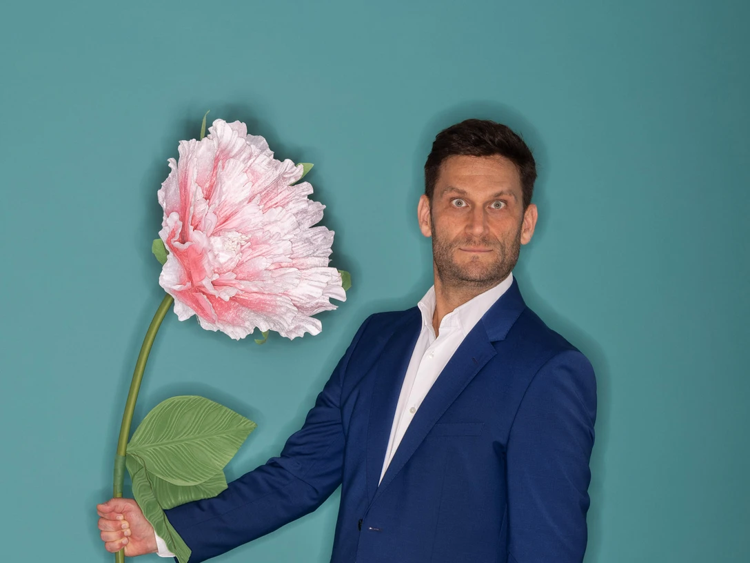 Pressefoto CH_Versöhnung_Fotograf Horst Klein_9.jpg Ein Mann in einem blauen Anzug hält eine übergroße rosa Blüte vor einem türkisfarbenen Hintergrund.