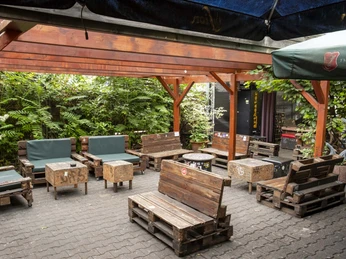 artheater Holzbänke und Tische aus Paletten unter einem Holzdach, umgeben von dichtem Grün.Wooden benches and tables made from pallets under a wooden roof, surrounded by dense greenery.