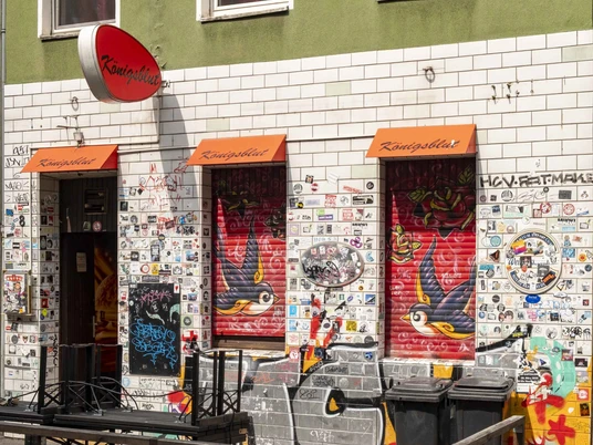 Königsblut Die Außenwand eines Lokals in Köln ist übersät mit Stickern und bunten Graffiti, an zwei roten Markisen hängt der Schriftzug "Königsblut".The outside wall of a pub in Cologne is covered in stickers and colorful graffiti, with the words "Königsblut" hanging from two red awnings.