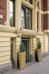 Die Fassade eines Gebäudes mit großen Fenstern und zwei modernen Pflanzgefäßen flankiert den Eingang.