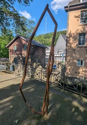Das Kunstwerk Dowan von Robert Schad auf den Skulpturenwegen Langenberg Skulptur Dowan von Robert Schad aus Cortenstahl, eingebettet in eine städtische Landschaft mit Fachwerkhäusern.