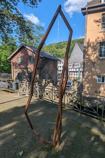 Das Kunstwerk Dowan von Robert Schad auf den Skulpturenwegen Langenberg Skulptur Dowan von Robert Schad aus Cortenstahl, eingebettet in eine städtische Landschaft mit Fachwerkhäusern.