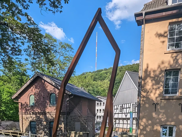 Das Kunstwerk Dowan von Robert Schad auf den Skulpturenwegen Langenberg Skulptur Dowan von Robert Schad aus Cortenstahl, eingebettet in eine städtische Landschaft mit Fachwerkhäusern.