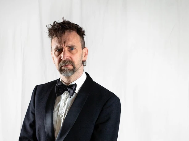 Iso Herquist Mann im Anzug neben einem Stuhl mit rosa Decke.