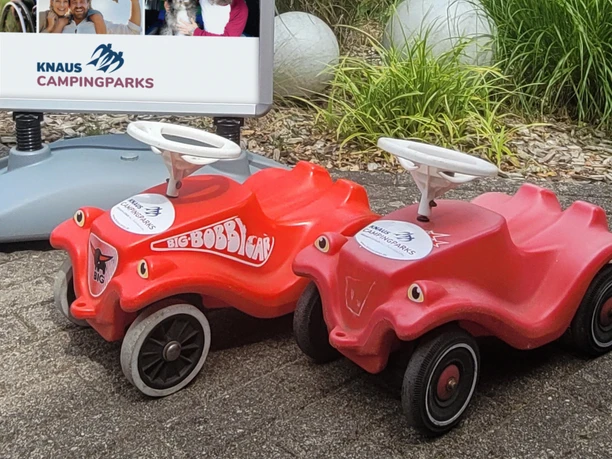 2_kcp_haren_Kidstime.jpg Zwei rote Kinderfahrzeuge vor einem Schild von Knaus Campingparks auf gepflastertem Boden.
