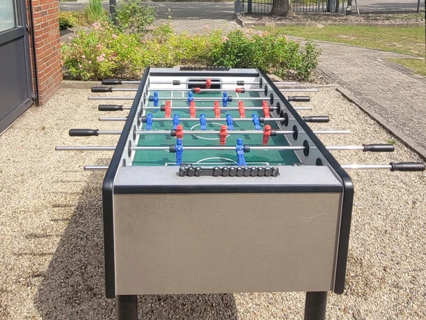 3a_kcp_haren_kicker_20210909_130629_schnitt.jpg Tischkicker mit blauen und roten Spielfiguren im Außenbereich auf Kiesfläche bei Sonnenschein.
