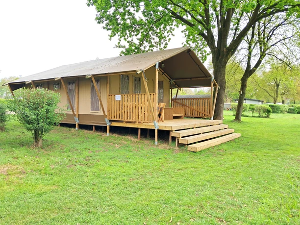 4_kcp_haren_safarizelt_serengeti1.jpg Safarizelt mit Holzterrasse auf grüner Wiese unter Bäumen im KNAUS Campingpark Haren