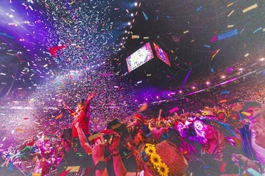 Lachende Kölnarena Inmitten eines glitzernden Konfettiregens und leuchtender Lichter feiert eine große Menschenmenge ausgelassen in der LANXESS arena. Bunte Blütenkränze und fröhliche Gesichter vor einer strahlenden Bühne zeugen von festlicher Stimmung und einem unvergesslichen Ereignis.