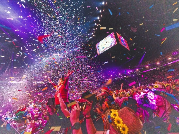 Lachende Kölnarena Inmitten eines glitzernden Konfettiregens und leuchtender Lichter feiert eine große Menschenmenge ausgelassen in der LANXESS arena. Bunte Blütenkränze und fröhliche Gesichter vor einer strahlenden Bühne zeugen von festlicher Stimmung und einem unvergesslichen Ereignis.