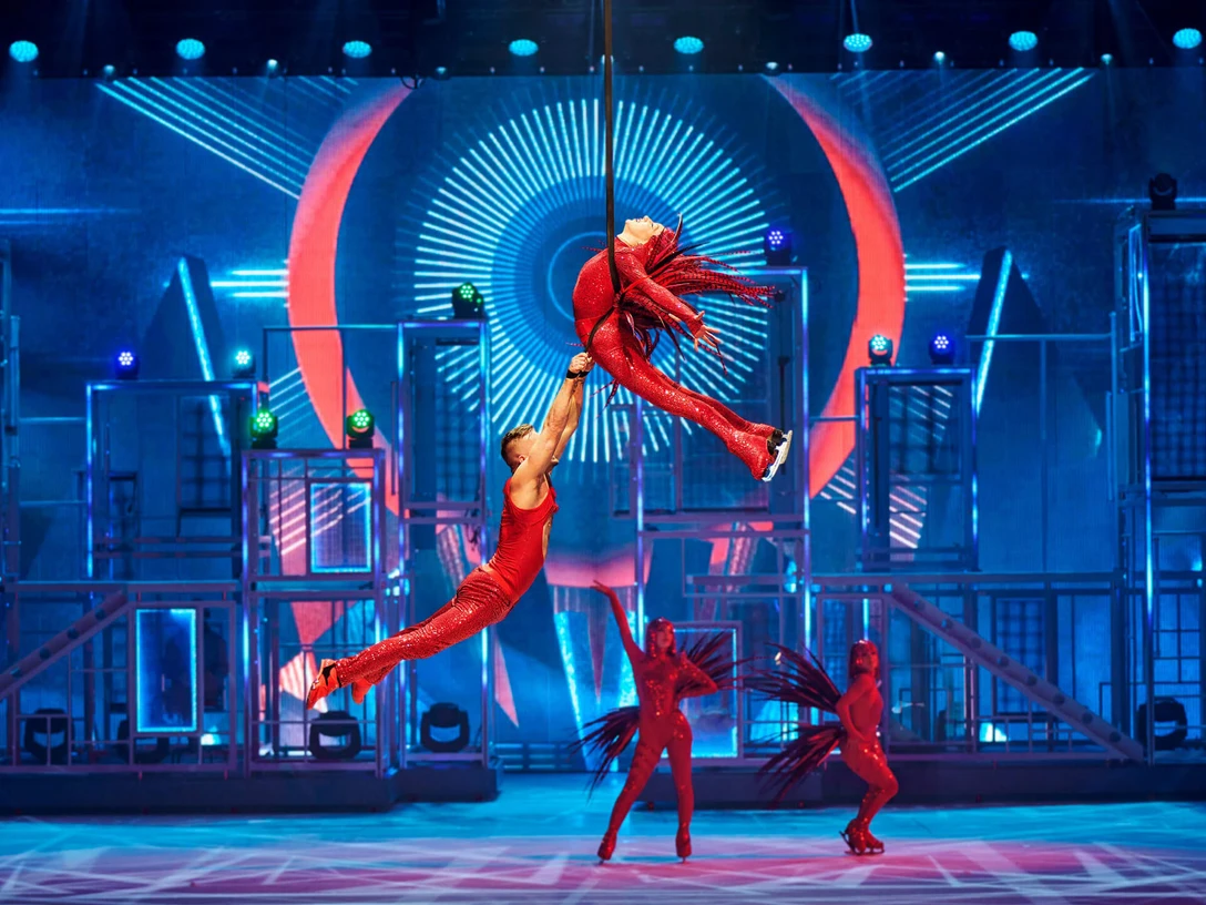 Akrobatische Eiskunstläuferinnen und -läufer in glitzernden roten Kostümen schweben elegant durch die Luft auf einer farbenfrohen Bühne mit neonbeleuchteten Hintergründen.Acrobatic figure skaters in glittering red costumes float elegantly through the air on a colorful stage with neon-lit backdrops.