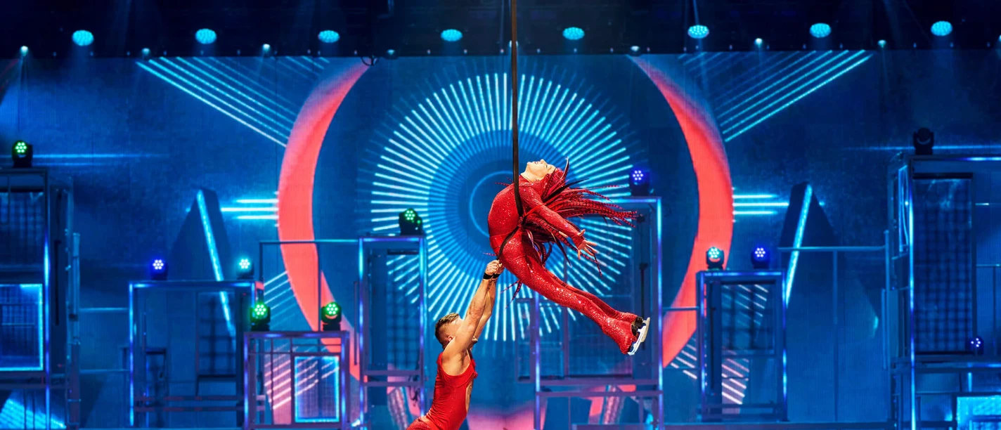 Acrobatic figure skaters in glittering red costumes float elegantly through the air on a colorful stage with neon-lit backdrops.
