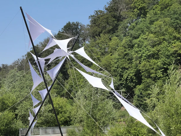 Das Kunstwerk Brückenschlag von Jens J. Meyer auf den Skulpturenwege Langenberg Weiße Segel aus Stoff, aufgespannt auf Metallstäben, stehen vor einem Wald am Skulpturenweg.