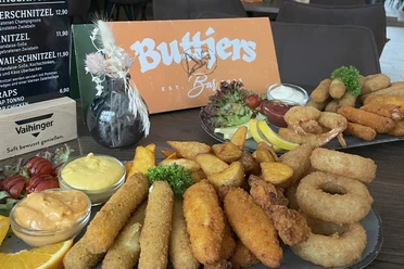 Buttjers Bar_1.jpg Eine Auswahl an frittierten Snacks und Saucen neben Gastrokarten, präsentiert auf einem Tisch.