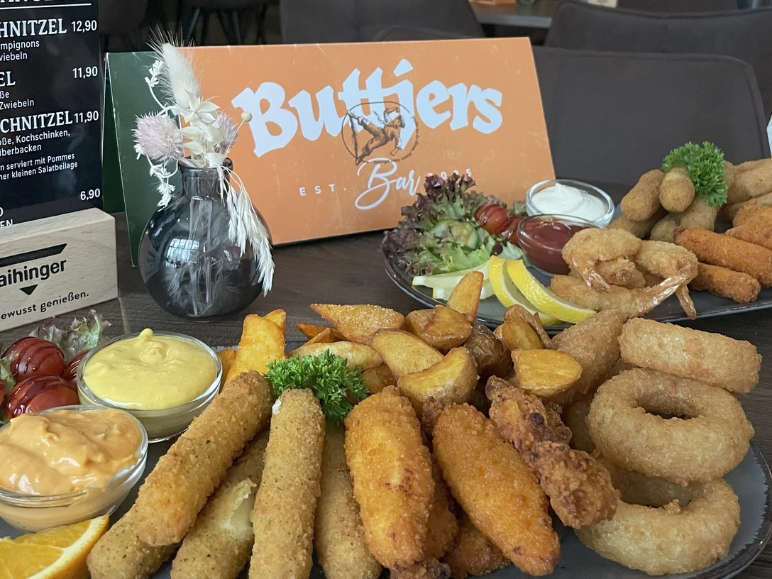 Buttjers Bar_1.jpg Eine Auswahl an frittierten Snacks und Saucen neben Gastrokarten, präsentiert auf einem Tisch.