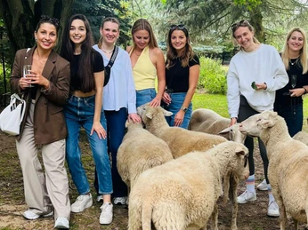 4.png Eine Gruppe von sieben lächelnden Frauen posiert fröhlich mit Schafen im Grünen.A group of seven smiling women happily pose with sheep in the countryside.