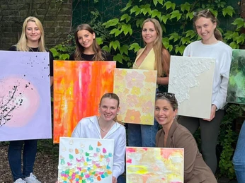 5.png Eine Gruppe junger Frauen präsentiert stolz ihre bunten, abstrakten Gemälde vor einer grünen Hecke.A group of young women proudly present their colorful, abstract paintings in front of a green hedge.