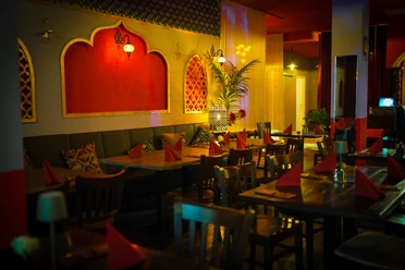 Mandaloun.jpg Innenansicht eines orientalischen Restaurants mit roten Wänden, Holzstühlen und weich beleuchteten Lampen.