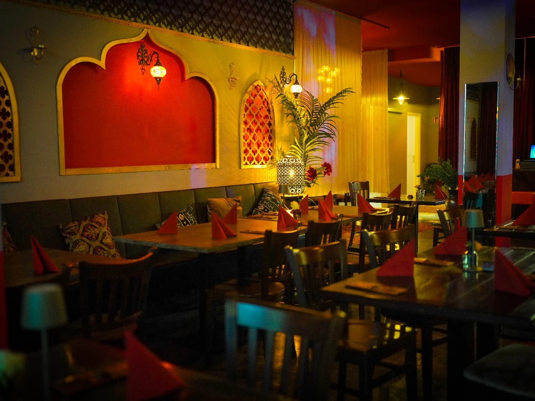Mandaloun.jpg Innenansicht eines orientalischen Restaurants mit roten Wänden, Holzstühlen und weich beleuchteten Lampen.