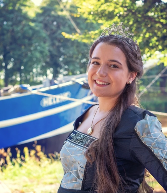 Lena Kiepe - amtierende Püntkerkönigin ©Heimatverein Haren (Ems) e.V..jpg Junge Frau in festlicher Tracht mit Krone lächelt vor einem blauen Boot im sonnigen Grünen.