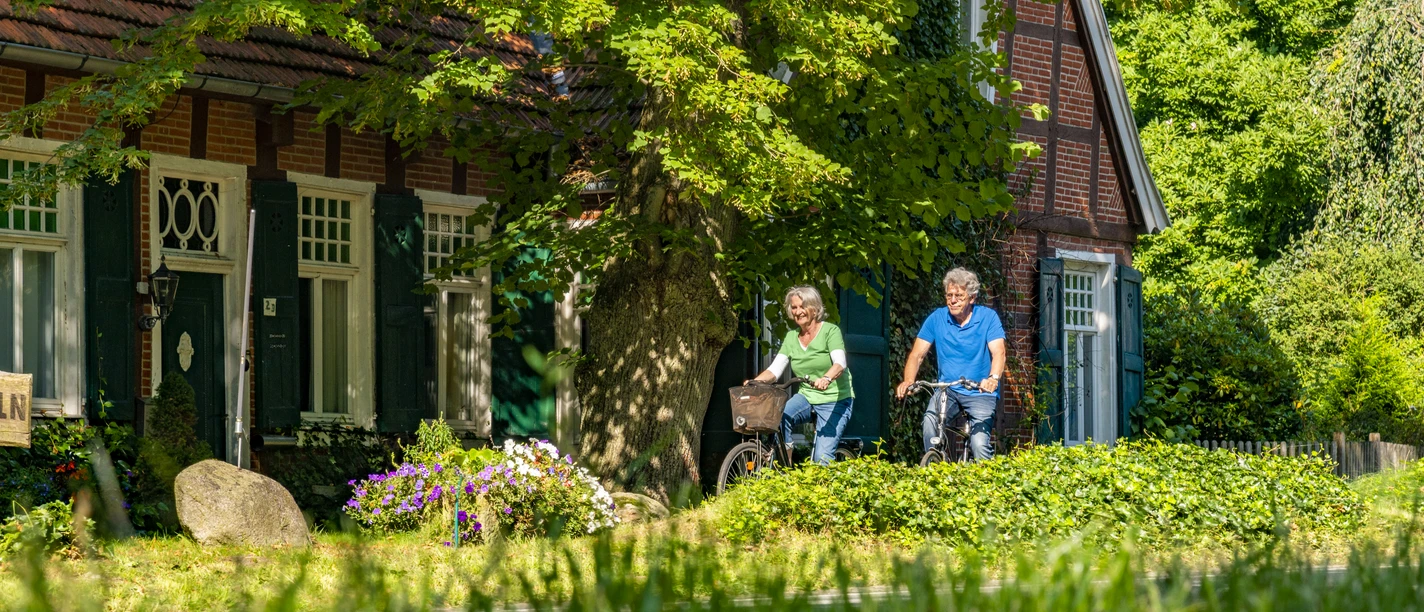 Hof Schroeter & Wielage, Bawinkel - Radfahren im Emsland ©Emsland Tourismus GmbH (1).jpg Zwei Radfahrer passieren ein rotes Backsteinhaus unter schattigen Bäumen im grünen Emsland.