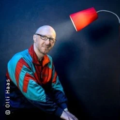 Ein Mann in bunter Jacke sitzt lächelnd vor dunkelblauem Hintergrund unter einer roten Lampe.