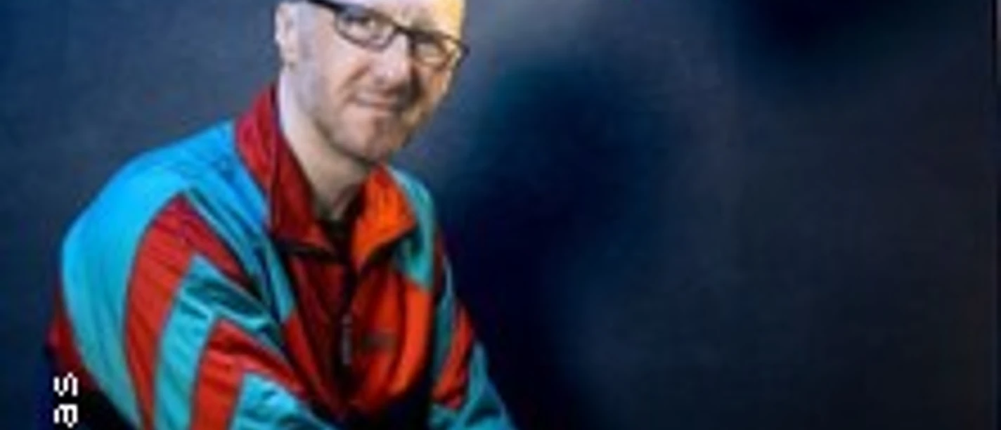 Ein Mann in bunter Jacke sitzt lächelnd vor dunkelblauem Hintergrund unter einer roten Lampe.
