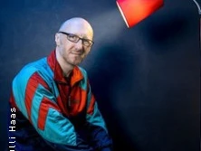 Ein Mann in bunter Jacke sitzt lächelnd vor dunkelblauem Hintergrund unter einer roten Lampe.