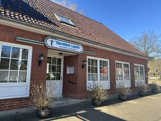 Taverne-Lakis von außen Backsteinbau mit weißen Fenstern, Eingangstür und Schild „Taverne Lakis – Griechische Spezialitäten“.