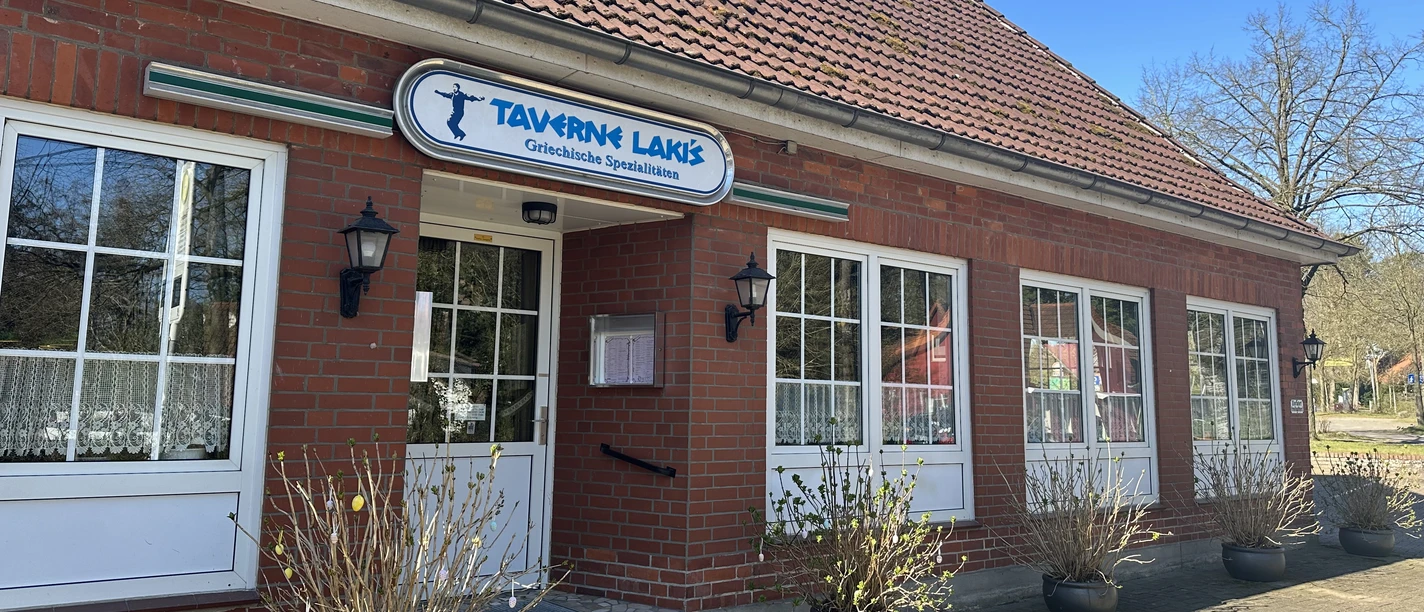 Taverne-Lakis von außen Backsteinbau mit weißen Fenstern, Eingangstür und Schild „Taverne Lakis – Griechische Spezialitäten“.