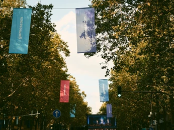 gamescom city festival Bunte gamescom-Fahnen hängen zwischen Bäumen, darunter ein belebter Straßenabschnitt.Colorful gamescom flags hang between trees, with a busy stretch of road below.