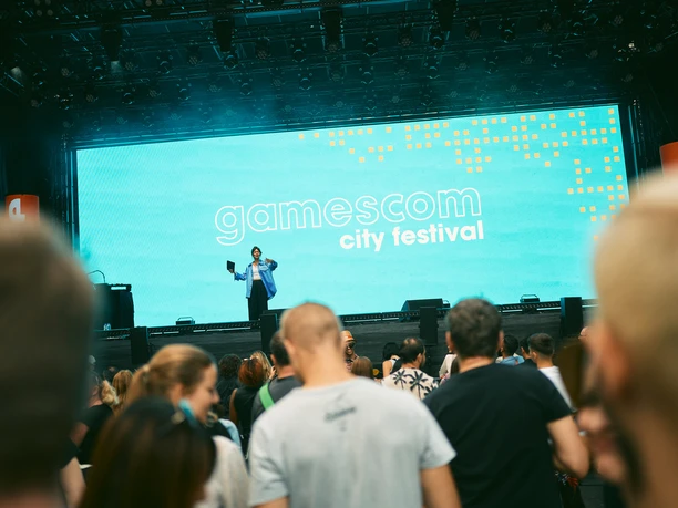 gamescom city festival A large crowd stands in front of a stage on which a male presenter is speaking. The "gamescom city festival" logo can be seen on the screen in the background.