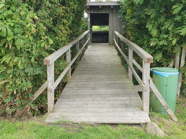 Vogelbeobachtungshütte Eine Vogelbeobachtungshütte aus Holz mit überdachtem Zugang und umgeben von grünen Büschen.