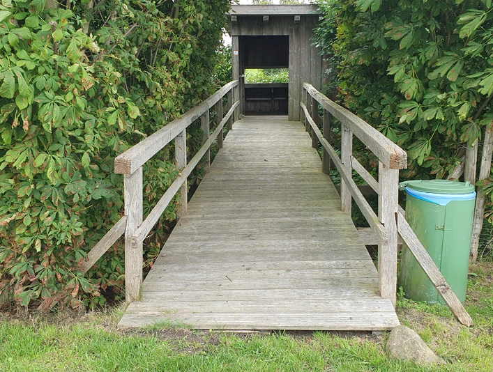 Vogelbeobachtungshütte Eine Vogelbeobachtungshütte aus Holz mit überdachtem Zugang und umgeben von grünen Büschen.