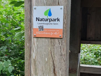 Infotafel Naturpark Lauenburgische Seen Ein hölzerner Pfosten trägt ein Schild des Naturparks Lauenburgische Seen. Der QR-Code gibt nähere Informationen zum Naturpark.