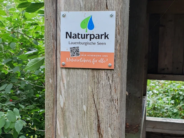 Infotafel Naturpark Lauenburgische Seen Ein hölzerner Pfosten trägt ein Schild des Naturparks Lauenburgische Seen. Der QR-Code gibt nähere Informationen zum Naturpark.
