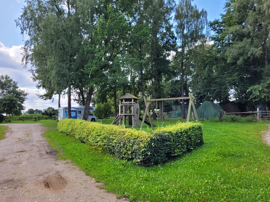 Spielplatz_in_Volkwardingen_.jpg pielplatz in Volkwardingen mit Kletterturm, Schaukel und Hecke, umgeben von Bäumen und Wiesen