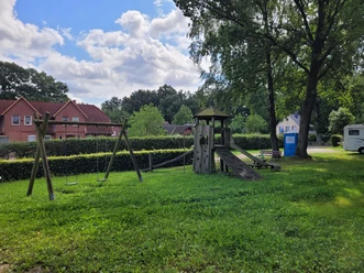 Spielplatz_in_Volkwardingen__01.jpg Spielplatz in Volkwardingen mit Schaukel, Kletterturm und Rutsche auf grünem Rasen, umgeben von Bäumen und Wohnhäusern