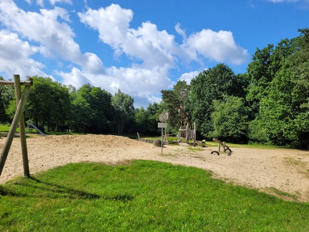 Badeteich_Steinbeck_.jpg Spielplatz mit Sandfläche, Klettergerüst und Schaukeln am Badeteich Steinbeck, umgeben von Bäumen unter blauem Himmel mit Wolken