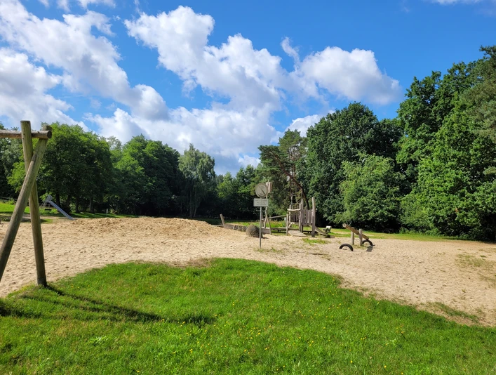 Badeteich_Steinbeck_.jpg Spielplatz mit Sandfläche, Klettergerüst und Schaukeln am Badeteich Steinbeck, umgeben von Bäumen unter blauem Himmel mit Wolken