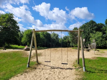 Badeteich_Steinbeck__01.jpg Zwei Holzschaukeln über Sandfläche im Spielbereich des Badeteichs Steinbeck, umgeben von Bäumen und blauem Himmel mit Wolken