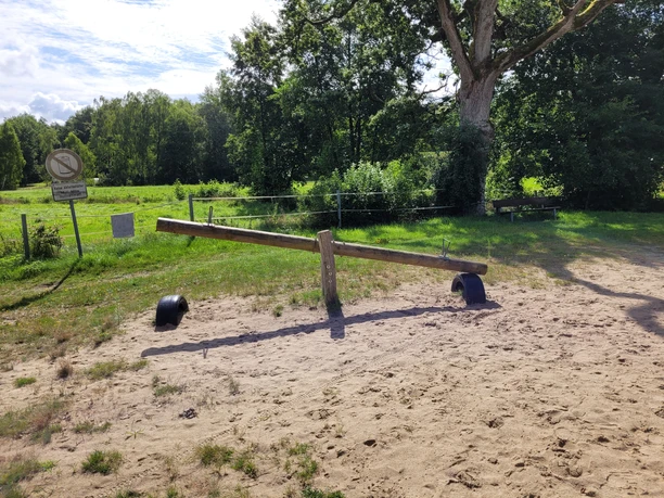 Badeteich_Steinbeck__02.jpg Holz-Wippe auf Sandfläche am Badeteich Steinbeck, umgeben von grünen Bäumen und Wiesen