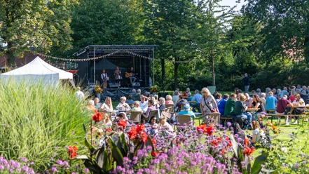 Open-air concert in the countryside with musicians on stage and audience at tables in the sunshine.