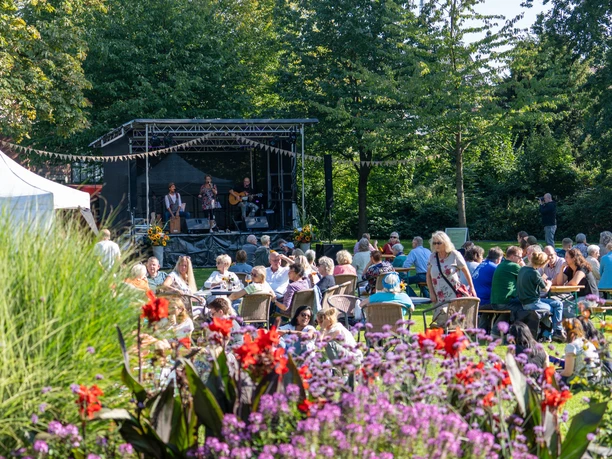 Spätsommerkonzert Open-Air-Konzert im Grünen mit Musikern auf Bühne und Publikum an Tischen im Sonnenschein.