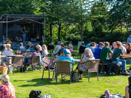 DSC00922.jpg Leute sitzen auf Stühlen im Freien, blicken auf eine Bühne mit Musikern.