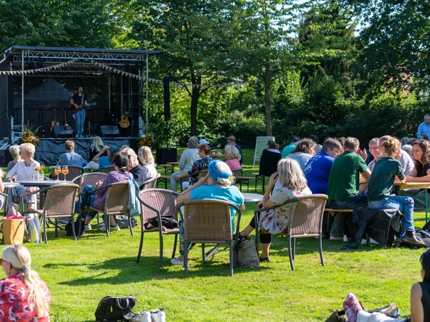 DSC00922.jpg People sit on chairs outside, looking at a stage with musicians.